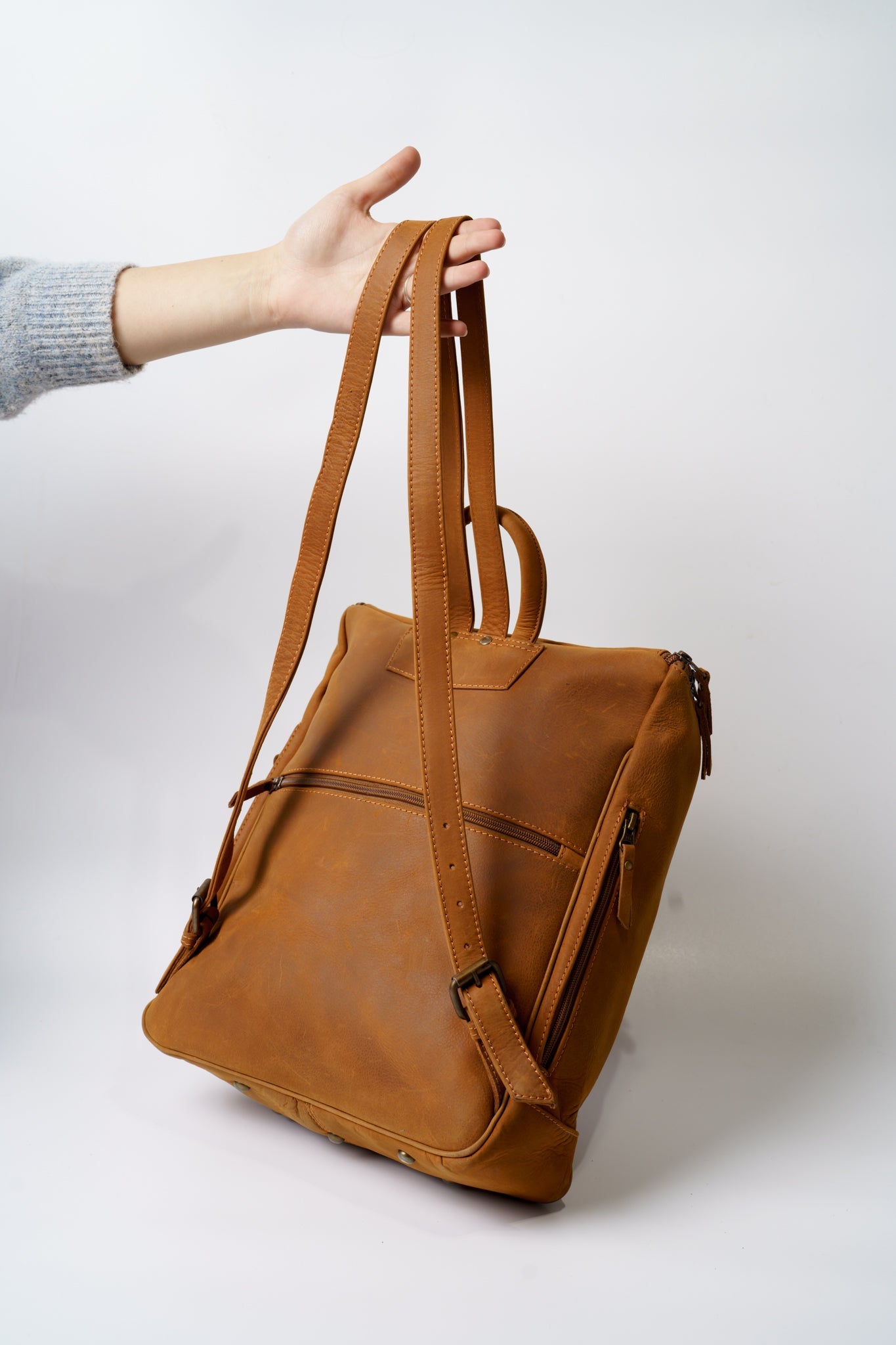 Rear view of Chamra's light brown satchel backpack. The bag can be seen dangling from the hand of a model, showcasing durability and the adjustable buckles on the shoulder straps.