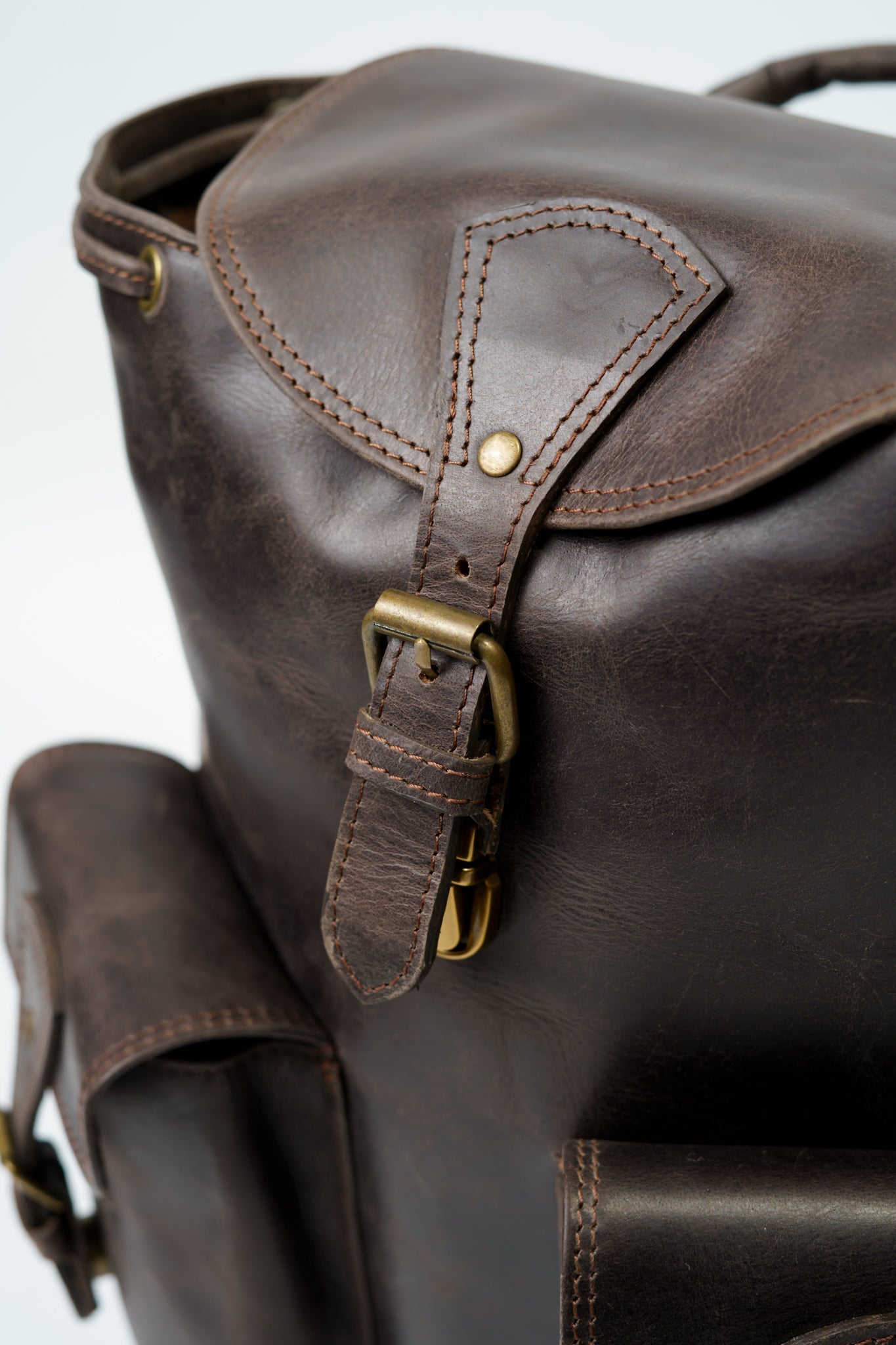 Close-up of the strap and buckle on the front pocket closure on Chamra's Brown Utility Leather Backpack. The image showcases high quality stitching, as well as the finish on the metal buckles.