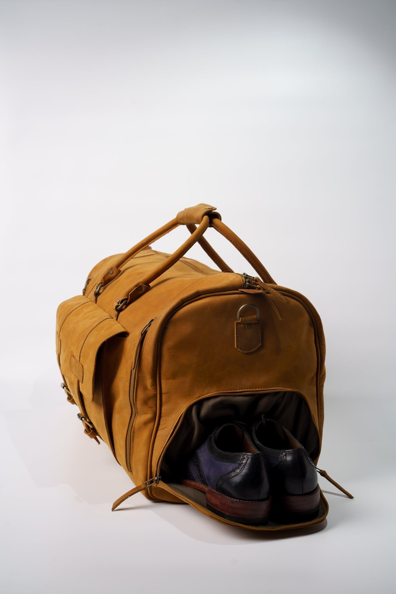 Side view of Chamra's Matte Leather Duffel bag with shoe compartment. This shot showcases the compartment for the shoes on the side of the bag. A pair of black oxford shoes can be seen popping out of the designated shoe compartment.