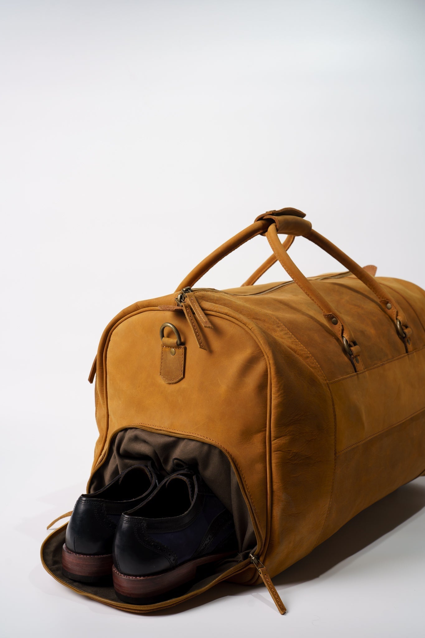 Side view of Chamra's Matte Leather Duffel bag with shoe compartment. This shot showcases the compartment for the shoes on the side of the bag. A pair of black oxford shoes can be seen popping out of the designated shoe compartment.